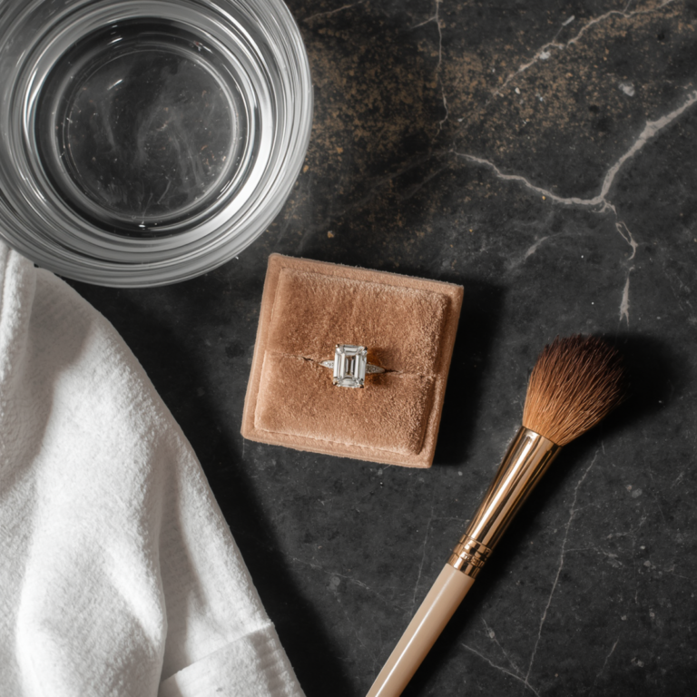 Flat lay of emerald-cut diamond ring on champagne velvet with glass water bowl, microfiber cloth, and gold makeup brush on dark marble
