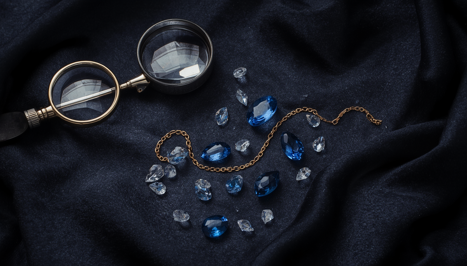 Flat-lay of jeweler's loupes with loose blue sapphires and clear topazes and gold-filled chain on dark textured silk with moody luxury lighting